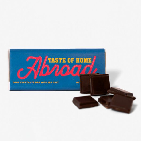 A dark chocolate bar with sea salt branded "taste of home abroad" displayed with broken pieces in front of its packaging.